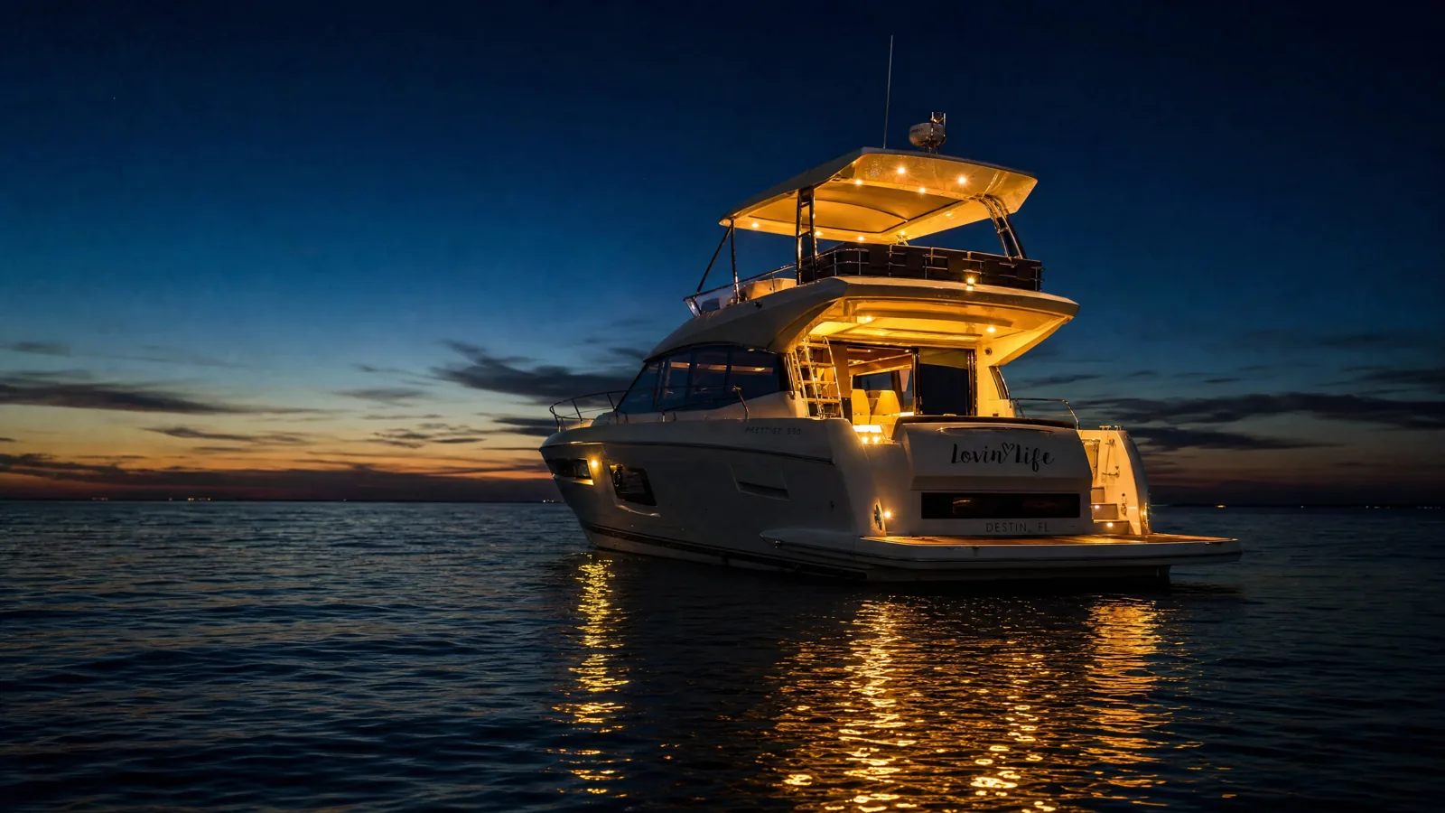 Private Evening Yacht Charter on the Emerald Coast at Dusk The Lovin’ Life yacht at anchor on calm Gulf water at dusk, deck lights glowing gold and reflecting across the surface as the last sunset colour fades from the horizon